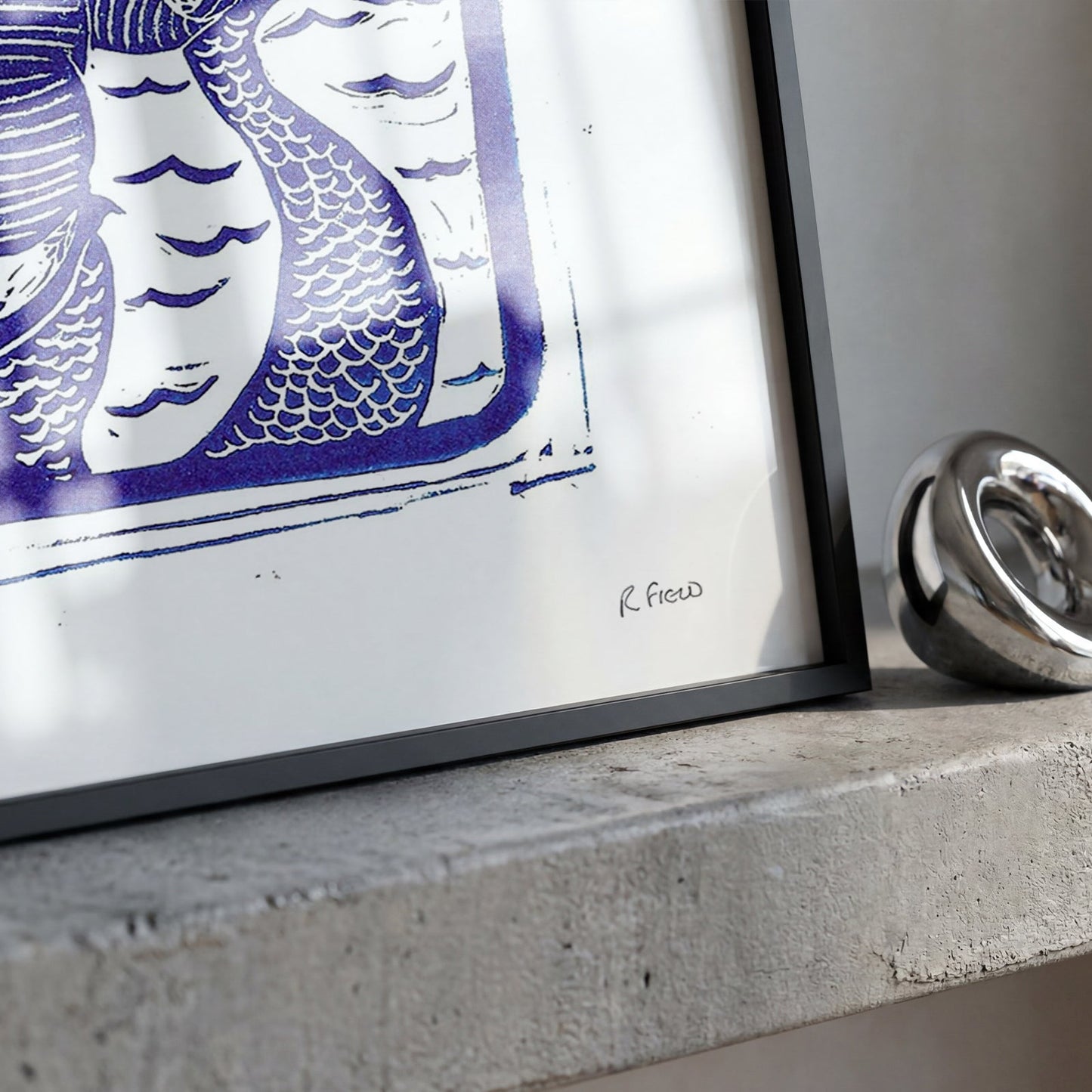 A framed art print featuring a stylized blue and white illustration of a merman, displayed on a textured concrete ledge next to a polished silver metallic ring.
