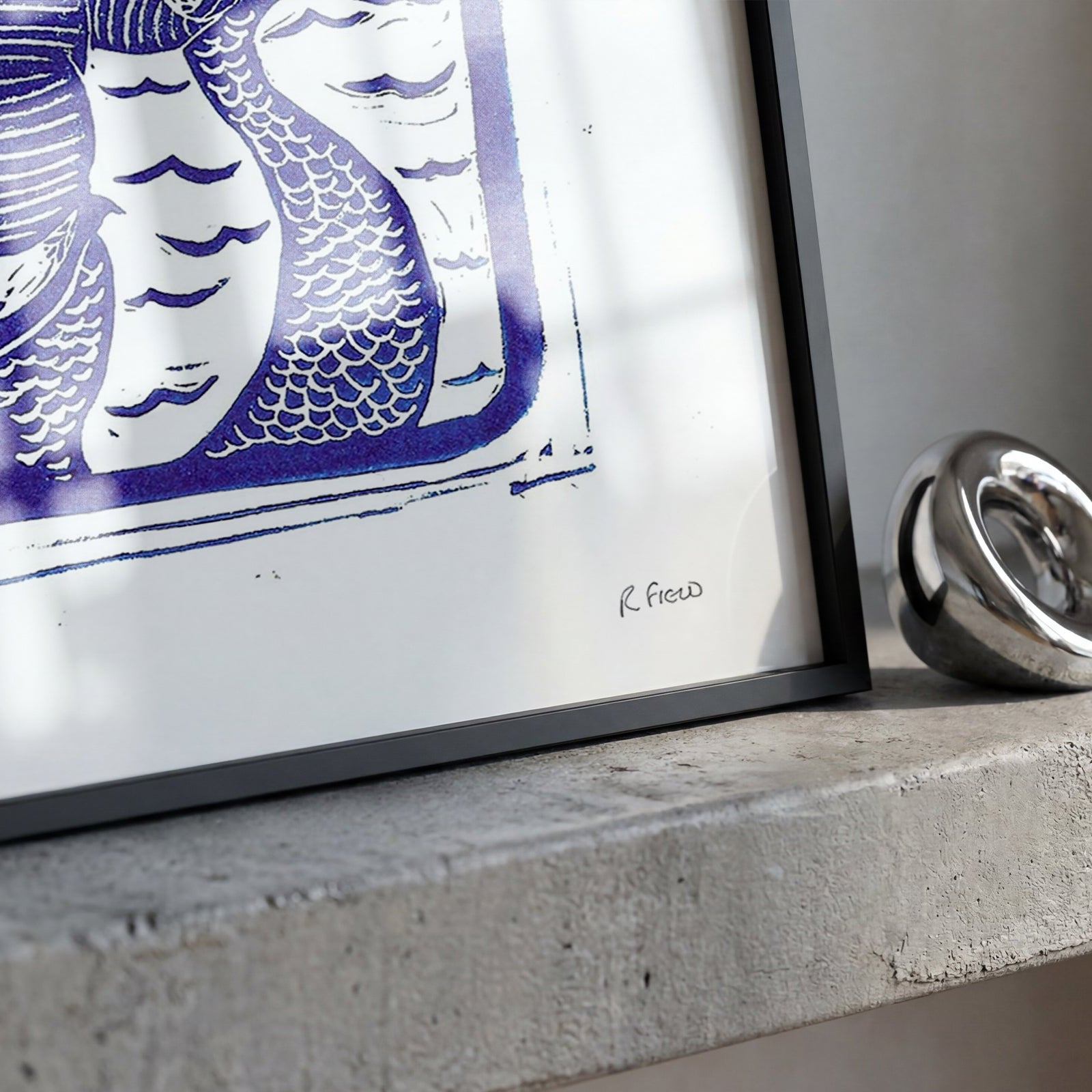 A framed art print featuring a stylized blue and white illustration of a merman, displayed on a textured concrete ledge next to a polished silver metallic ring.