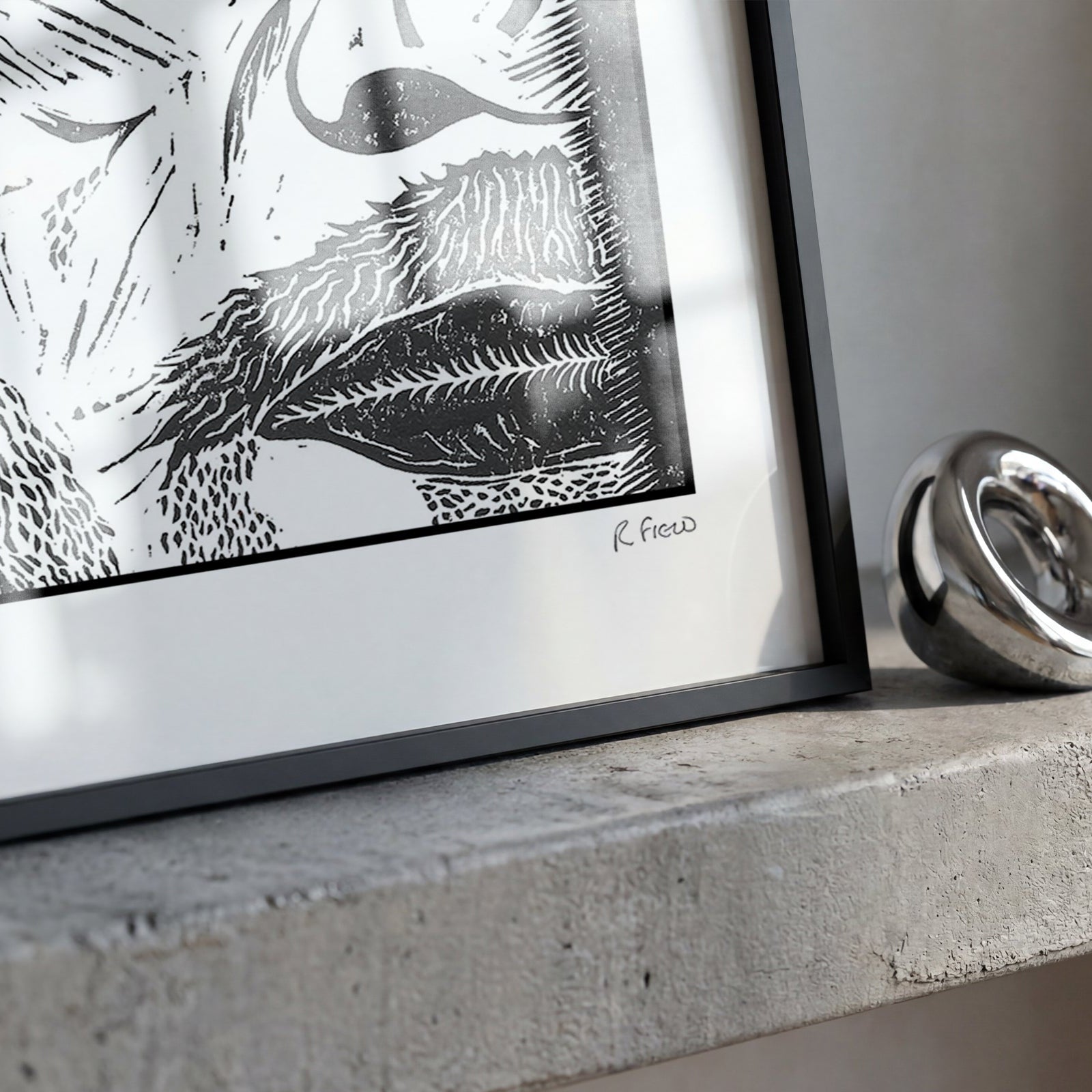 A framed black and white art print with a black frame and white mat, resting on a concrete ledge next to a polished silver metal ring.