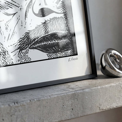 A framed black and white art print with a black frame and white mat, resting on a concrete ledge next to a polished silver metal ring.