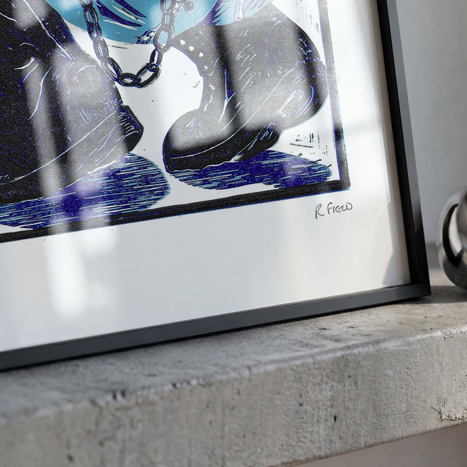A framed art print with a black frame and white mat, featuring a graphic design in blue, black, and white, resting on a concrete ledge next to a shiny silver ring sculpture.