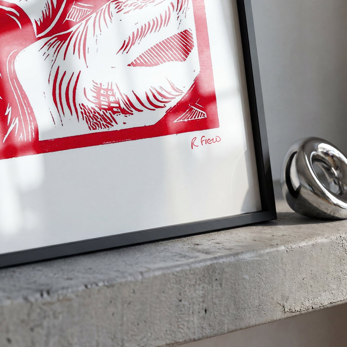 A framed red and white art print with a black frame leaning on a concrete ledge next to a shiny silver ring.