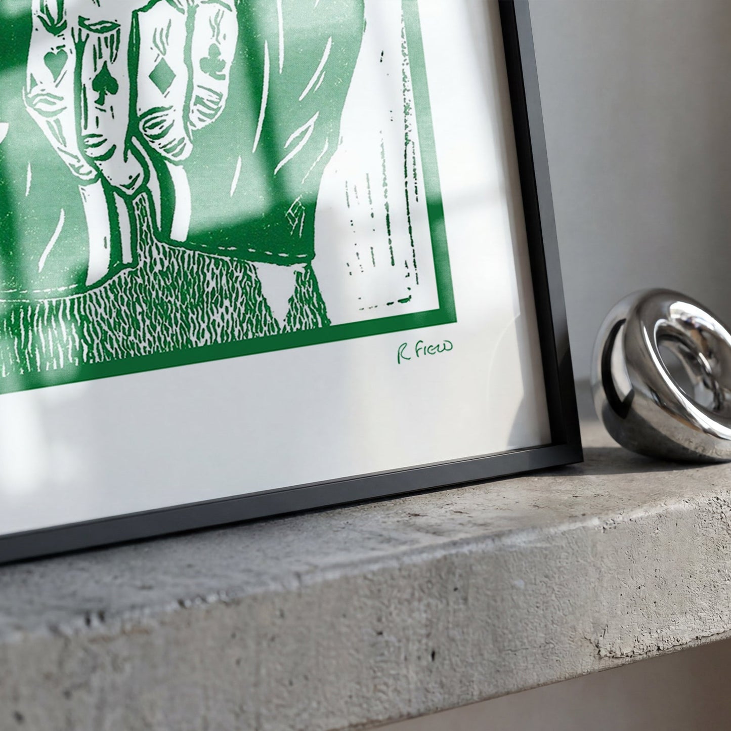 A framed green and white art print with a black frame, resting on a textured concrete ledge next to a shiny silver ring.