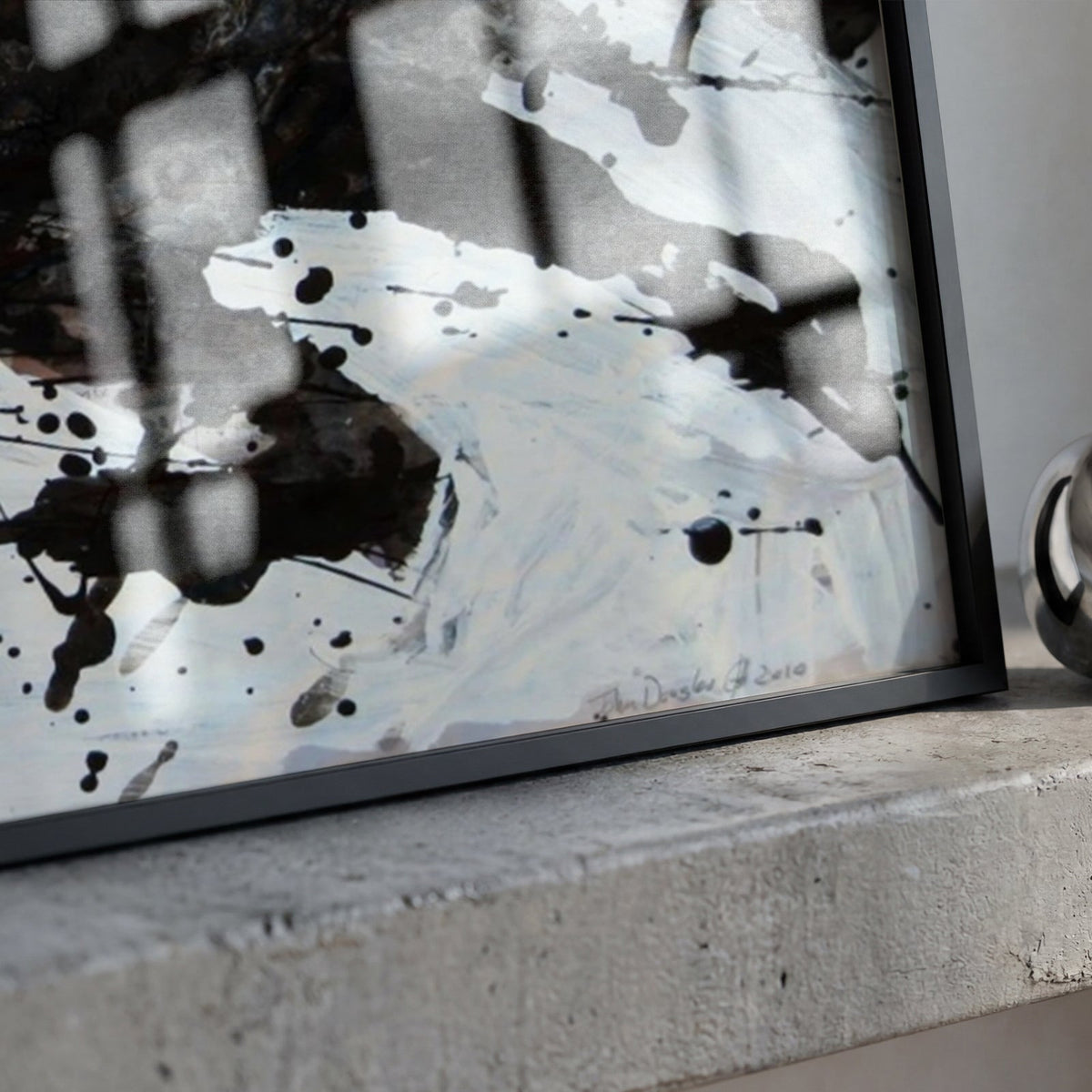 A framed abstract black and white print leaning against a concrete surface, with a reflective silver ring-shaped object beside it.