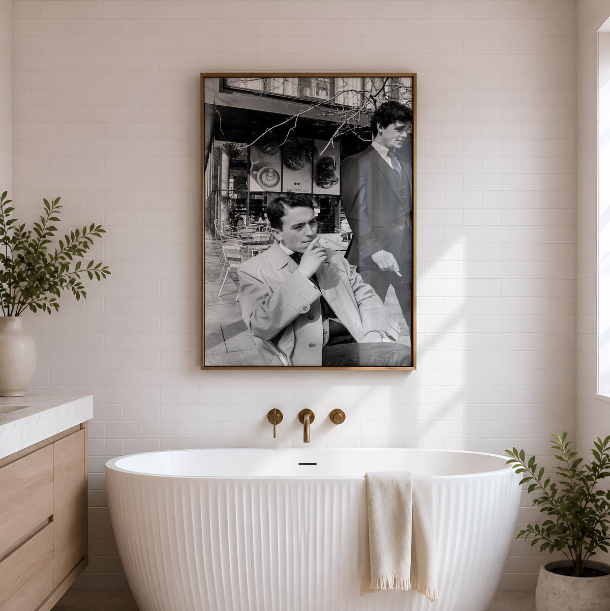 A freestanding ribbed white bathtub centered in a bright bathroom, with a large framed black-and-white portrait hanging on the wall above it. A folded cream towel rests on the tub, and potted plants sit on both sides.
