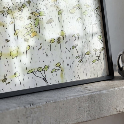 A framed art print featuring a delicate hand-drawn pattern of small green plants on a white background, resting on a textured gray concrete surface next to a polished silver metallic ring.