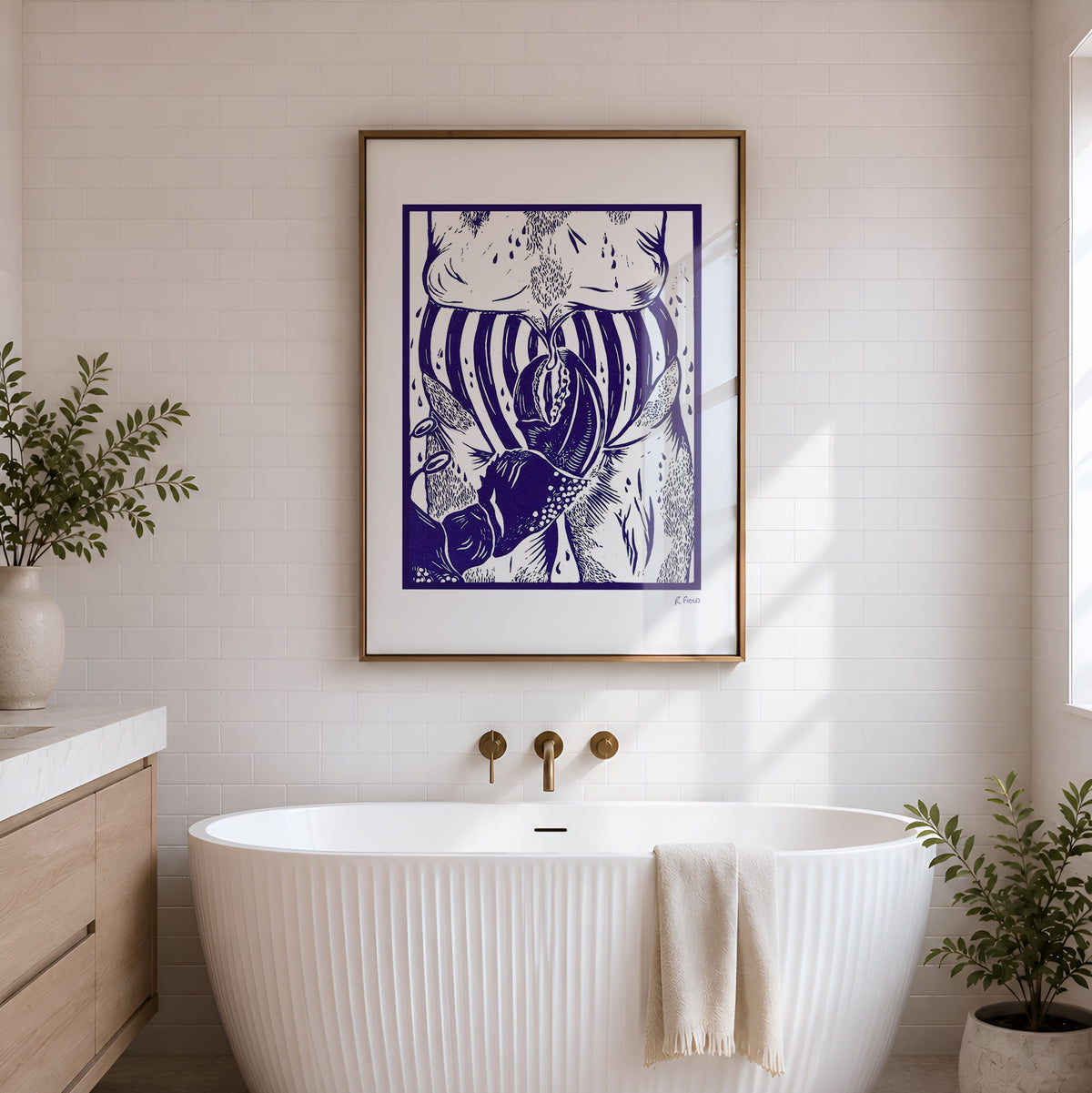 A modern freestanding bathtub in a bright bathroom with a framed blue and white linocut artwork hanging above it, plus potted plants and a wood vanity.