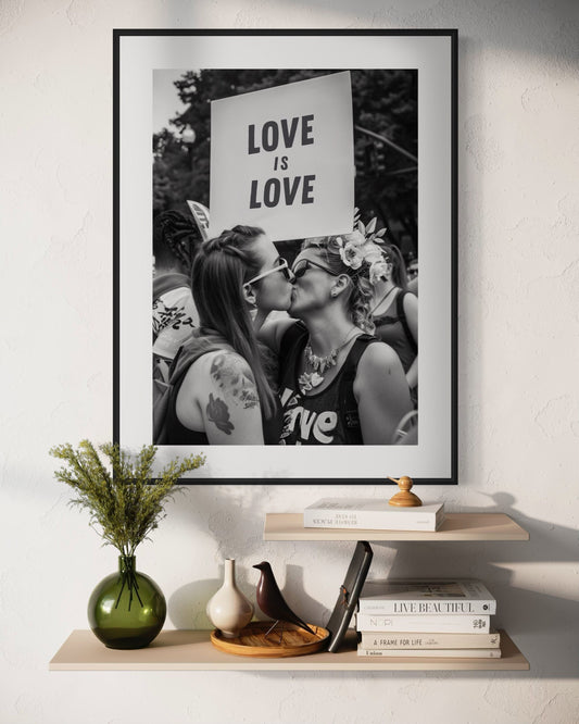 Black and white photograph of two women kissing at a Pride parade, with a sign behind them reading &#39;LOVE IS LOVE,&#39; symbolising equality and love in all forms.