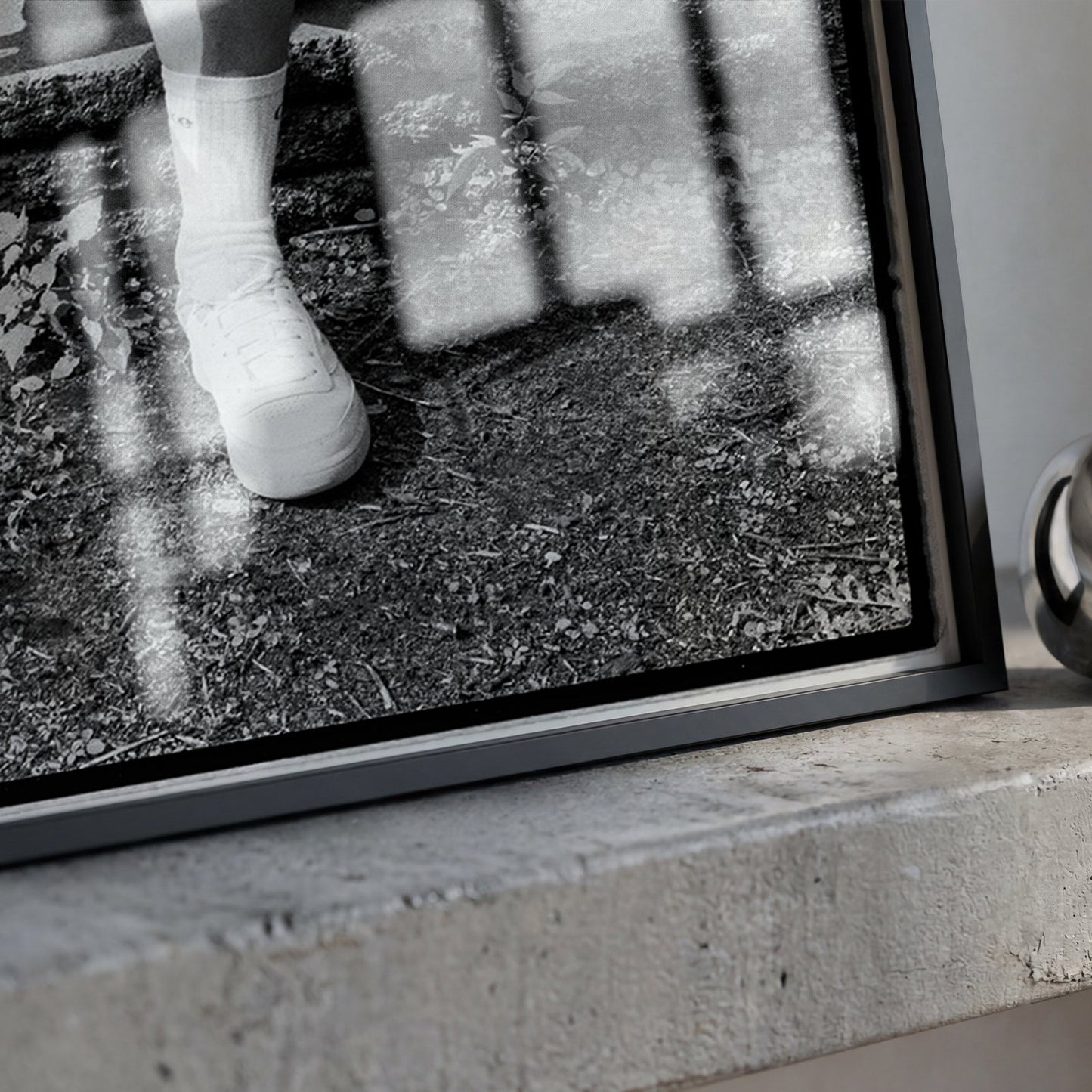 A framed black and white photograph leaning on a concrete ledge, showing a close-up of a white shoe and sock, next to a polished metallic ring-shaped object.