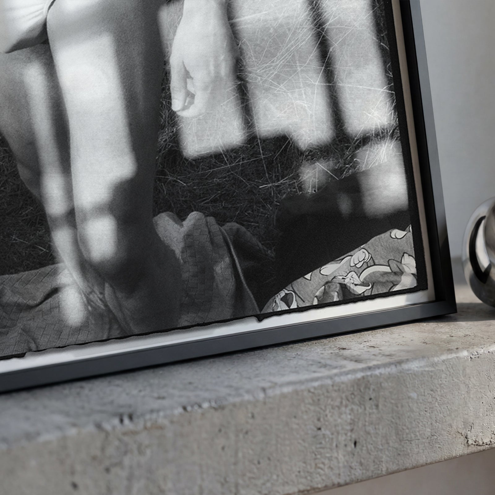 A black-framed black and white art print leaning against a concrete ledge, next to a polished silver metallic ring-shaped object.
