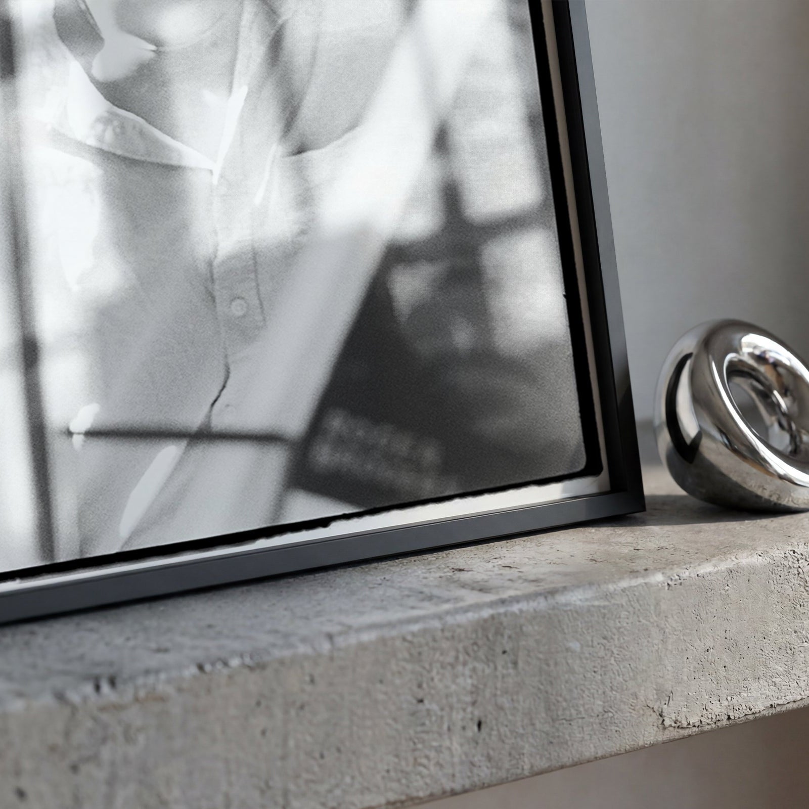A black-framed mirror leaning on a concrete ledge next to a polished silver metallic ring-shaped object.