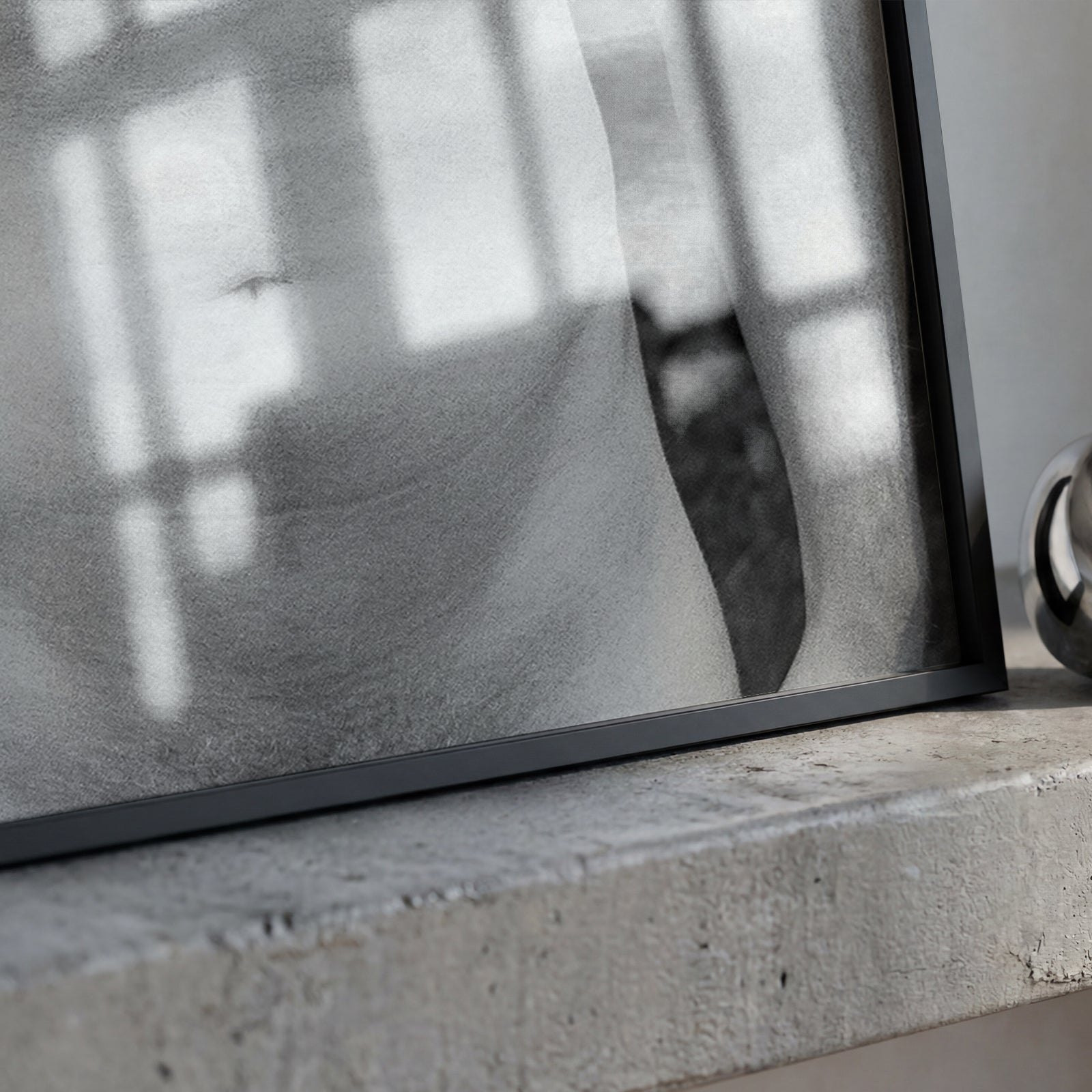 A black-framed art print with a black and white photograph of a draped figure, resting on a textured concrete ledge next to a shiny silver metallic ring sculpture.