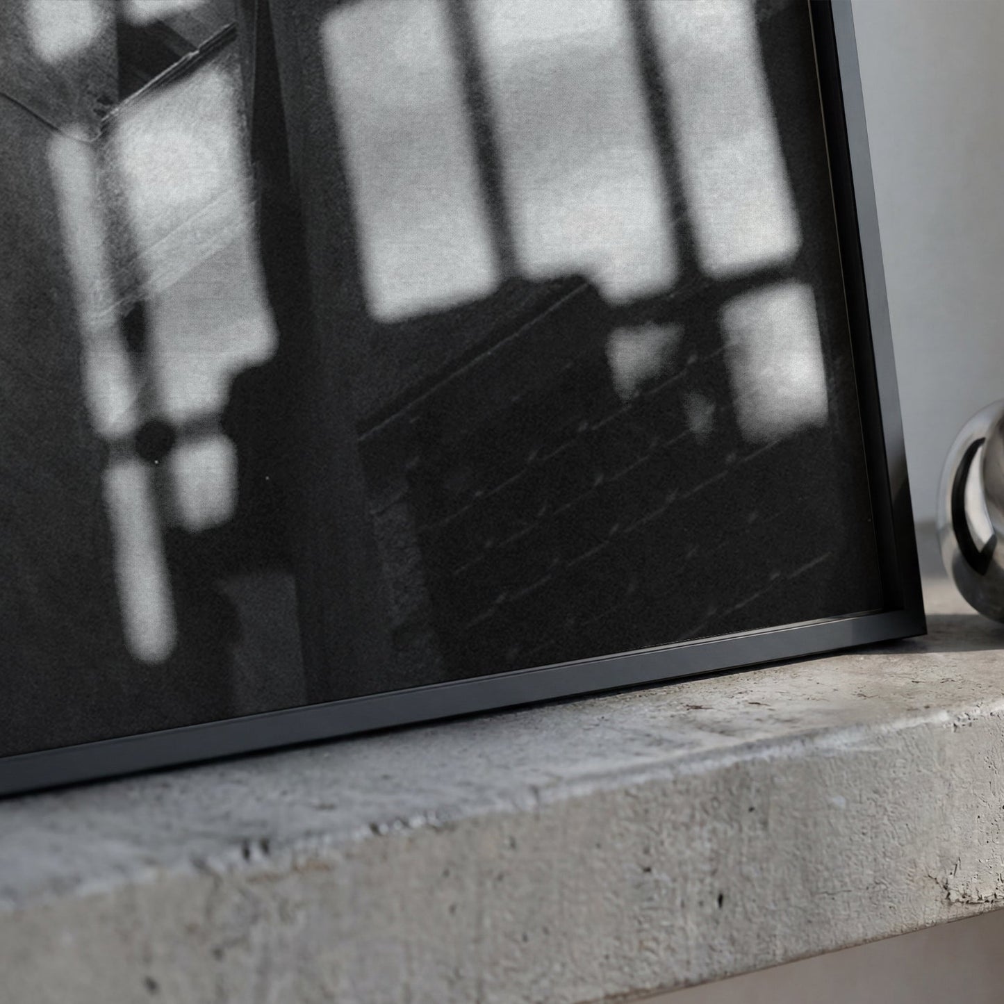 A black-framed art print with a black-and-white window shadow design, resting on a textured concrete ledge next to a polished silver metallic ring.